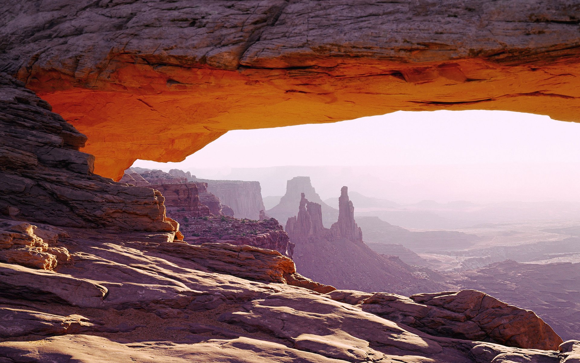HD PC desktop wallpaper capturing a natural rock arch framing a desert landscape with distant rock formations under a soft, hazy sky.