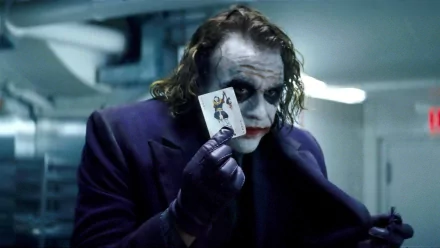 HD desktop wallpaper of the Joker from The Dark Knight movie, featuring the character in signature makeup and purple suit, holding a joker card close to his face in a dimly lit background.