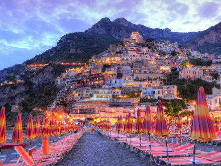 HD desktop wallpaper of Amalfi, Italy: colorful beach umbrellas on a Mediterranean shore leading to a lit, man-made terraced seaside town climbing the hillside at dusk.