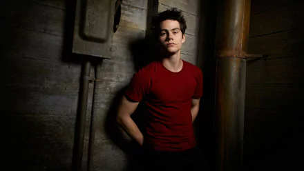 HD desktop wallpaper featuring Dylan O'Brien in a red T-shirt, leaning against a wall in a dimly lit setting.