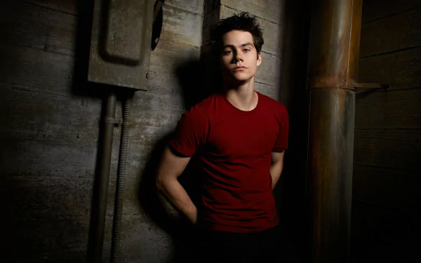 HD desktop wallpaper featuring Dylan O'Brien in a red T-shirt, leaning against a wall in a dimly lit setting.