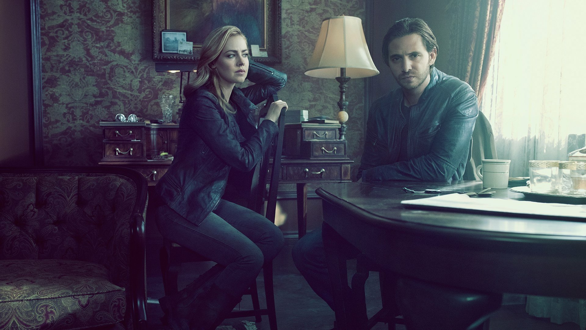 Two characters from the TV show 12 Monkeys sit at a dim vintage parlor table under a lamp, moody teal tones. HD PC desktop wallpaper and background.