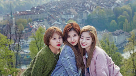 Three members of the music group TWICE pose together outdoors with a scenic background of a town. This HD desktop wallpaper and background captures their close bond.