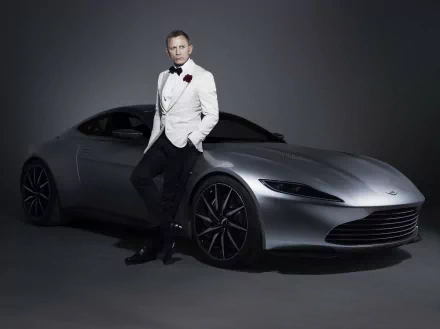 Man in a white tuxedo leaning against a silver Aston Martin under moody studio lighting, 007/James Bond–style HD desktop wallpaper.