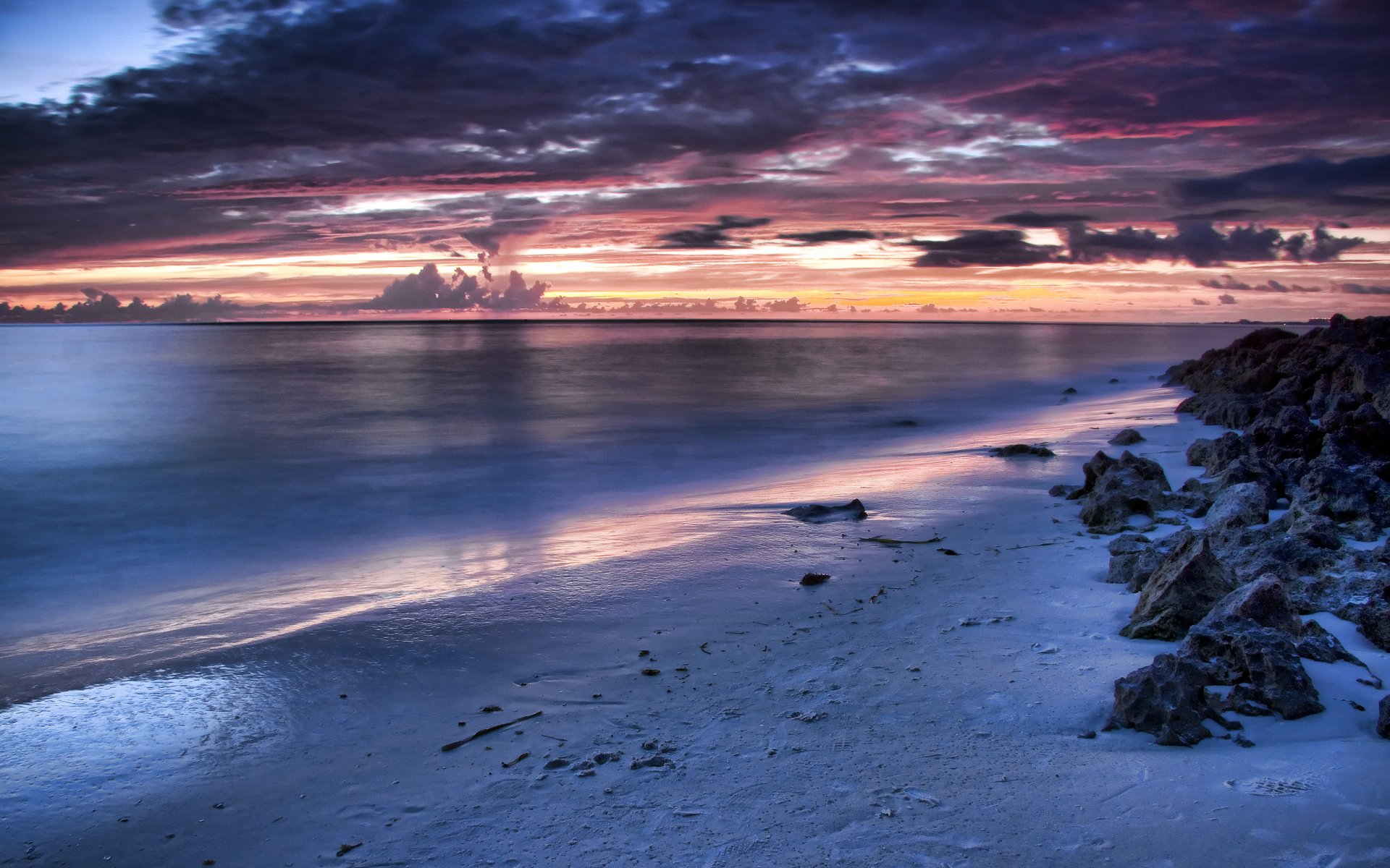 HD desktop wallpaper showing a tranquil beach at sunset with clouds reflecting vibrant colors over calm water and rocky shoreline.