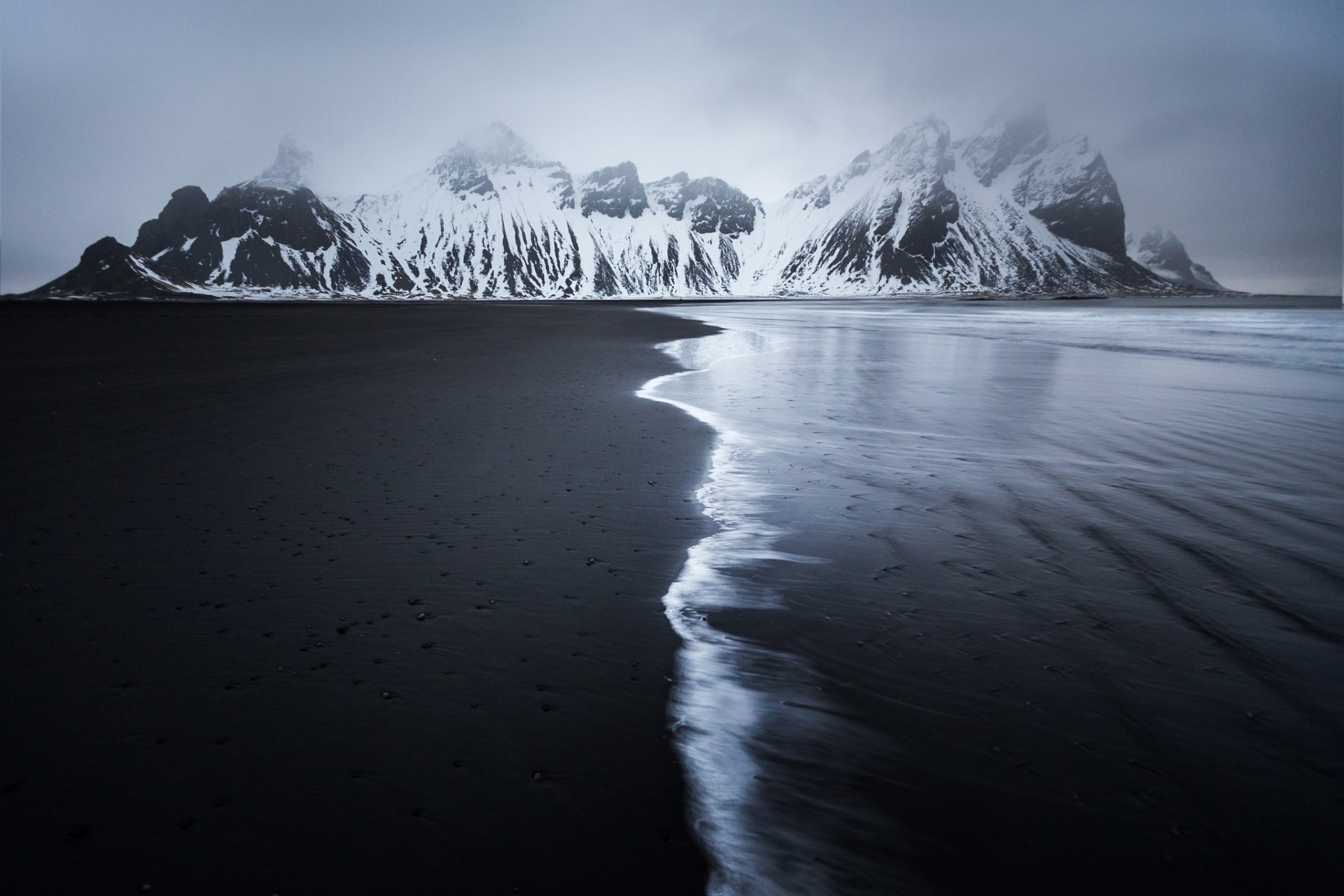HD desktop wallpaper of Vestrahorn mountains enveloped in fog, with dark sand and the ocean's edge creating a serene nature scene.