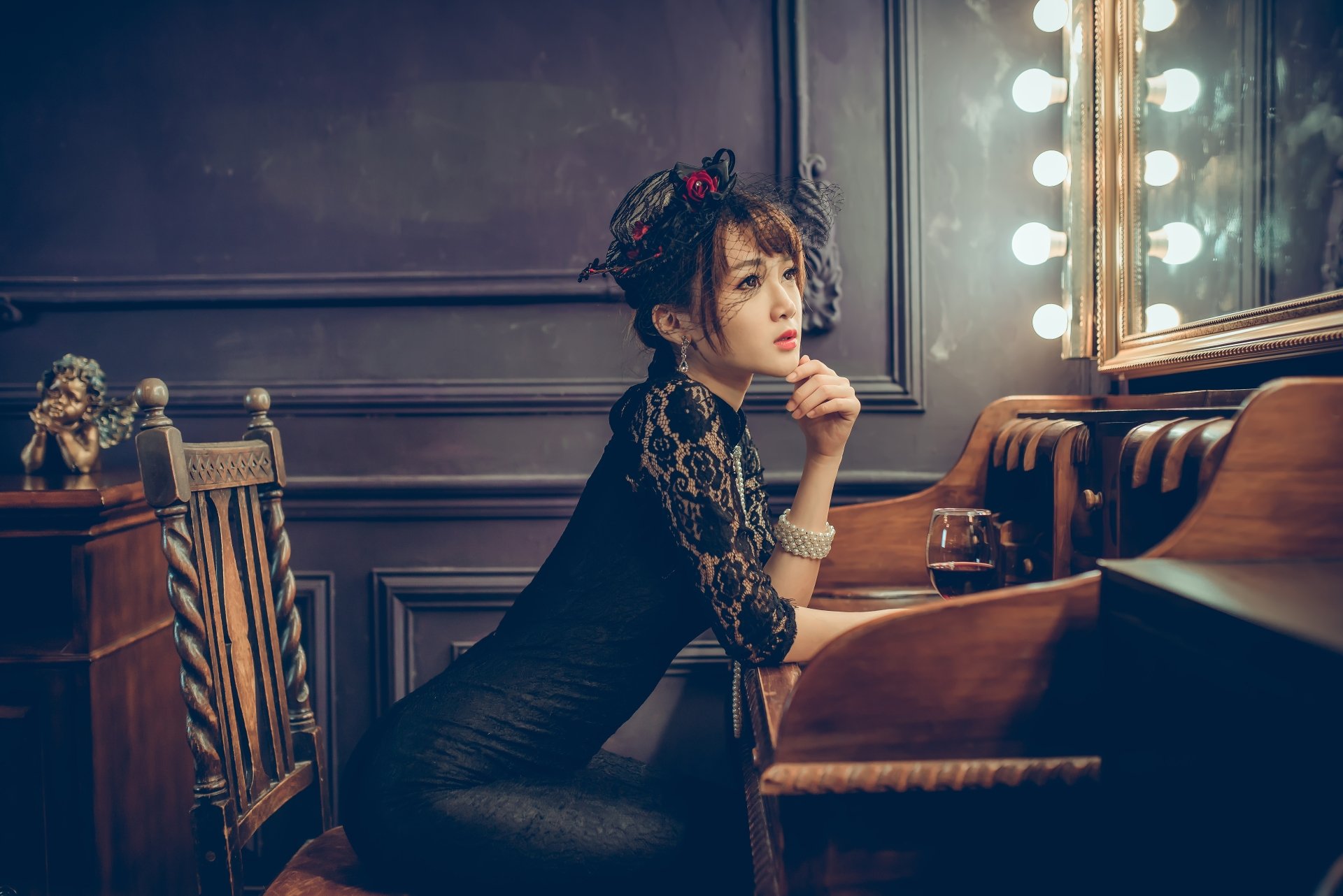 An Asian woman with brunette hair in a stylish dress and hat, posing thoughtfully at a vanity with a glass of wine in a moody, elegant setting. HD desktop wallpaper.