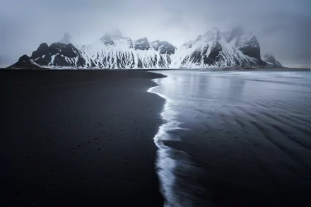 HD desktop wallpaper of Vestrahorn mountains enveloped in fog, with dark sand and the ocean's edge creating a serene nature scene.