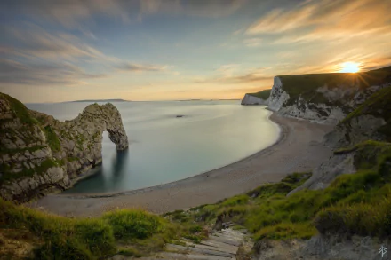 Sunset over Durdle Door, a natural limestone arch on the English coastline, with a serene ocean horizon and sandy beach embraced by green cliffs.