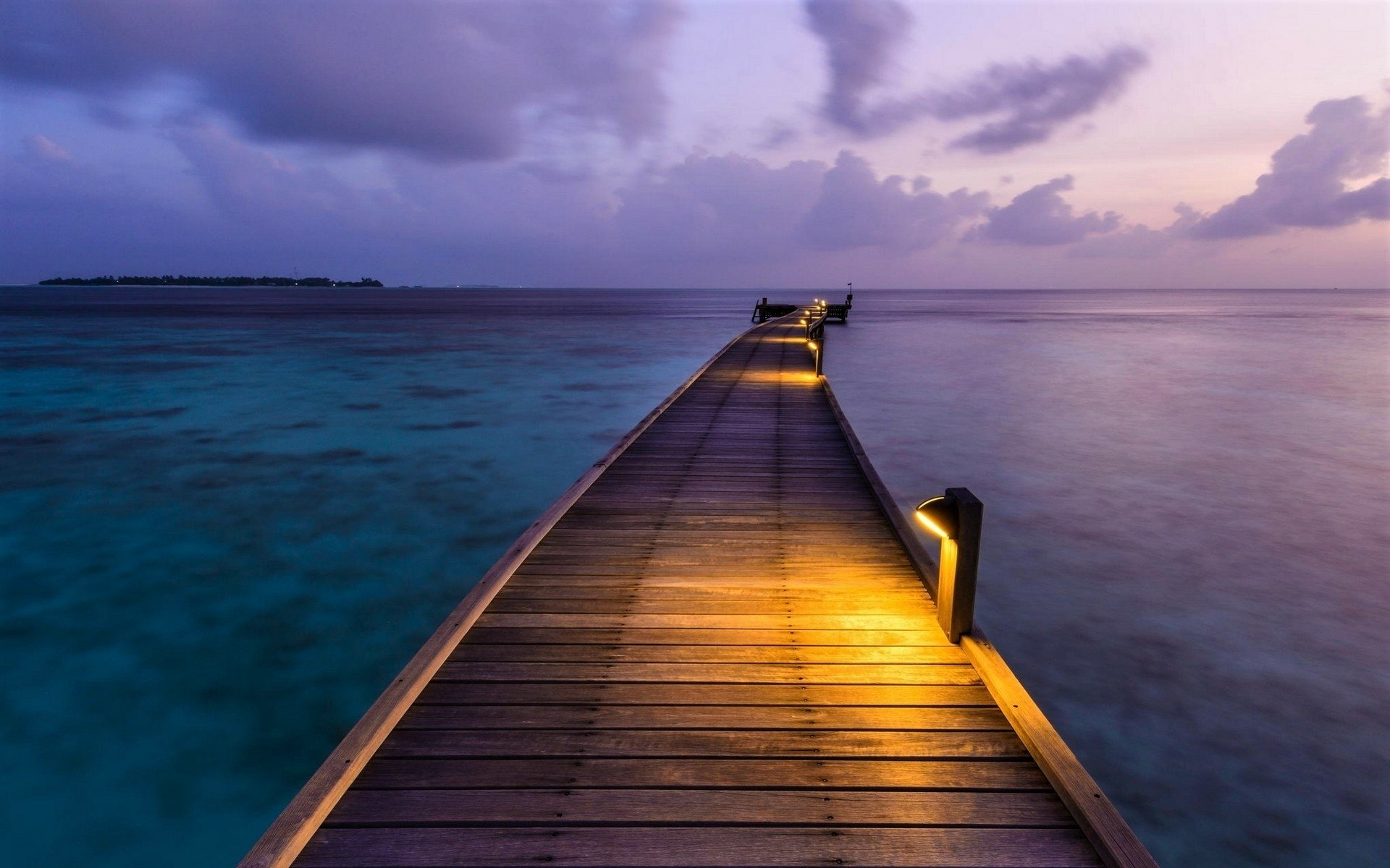 A wooden man-made pier extends over the calm ocean toward the horizon in the Maldives, illuminated by soft yellow lights under a purple twilight sky.