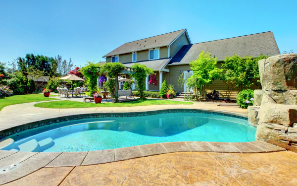 4K Ultra HD PC desktop wallpaper/background of a man-made backyard pool with rock feature, patio seating and lush landscaping in front of a two-story house.
