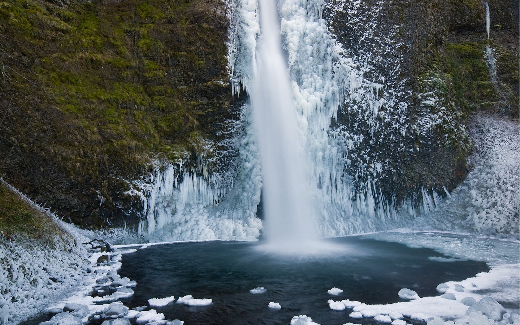 A stunning HD desktop wallpaper featuring a majestic waterfall cascading down icy cliffs into a serene pool below, surrounded by lush green moss and natural rock formations.