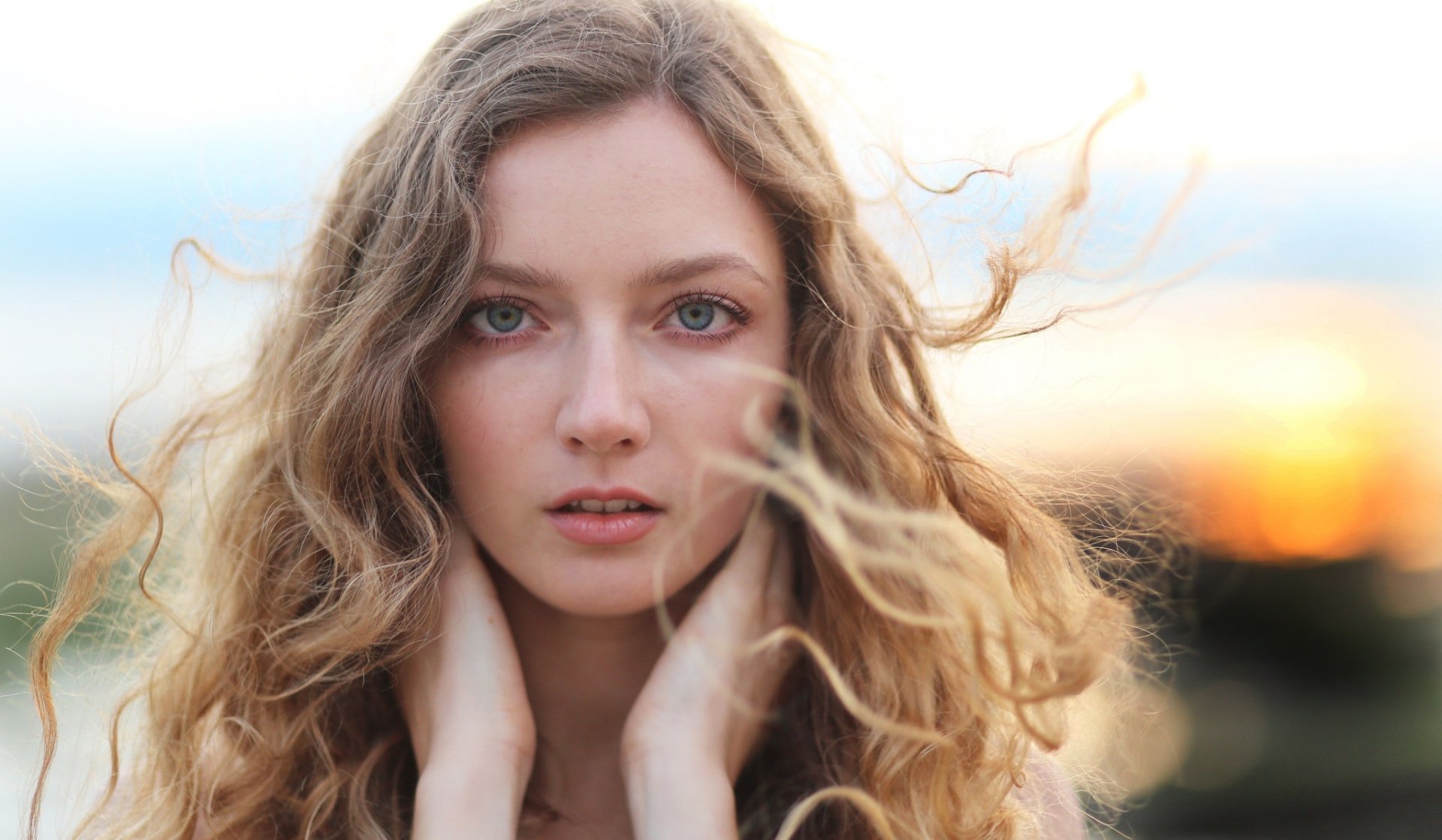 Close-up HD desktop wallpaper of a blonde woman with blue eyes, featuring a shallow depth of field that softly blurs the background at sunset.