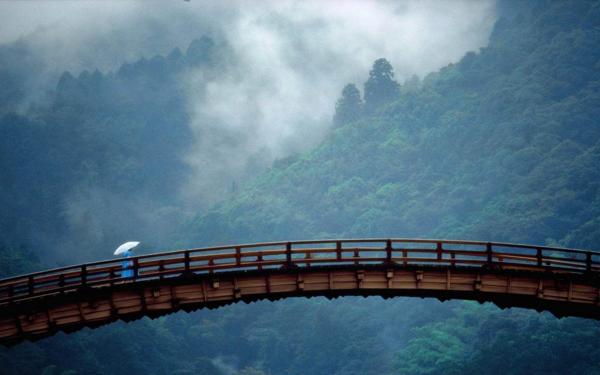 HD desktop wallpaper featuring a man-made wooden bridge with a lone figure holding a blue umbrella, set against a misty, forested mountain backdrop.