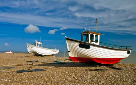 HD desktop wallpaper showing two boats resting on a pebble beach under a partly cloudy sky.