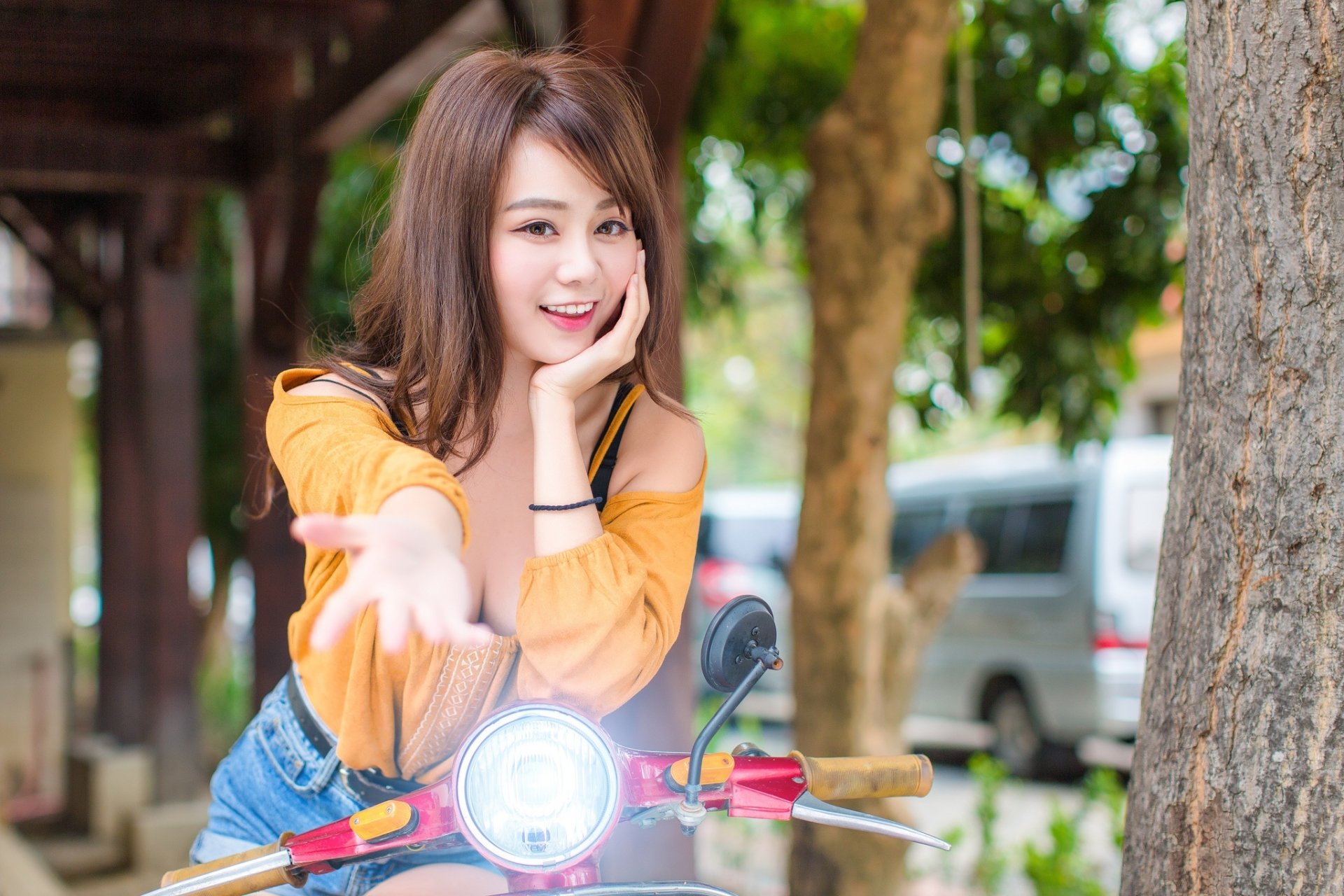 HD desktop wallpaper with a smiling brunette Asian woman, showing brown eyes, natural depth of field, posing casually by a tree with a blurred background.