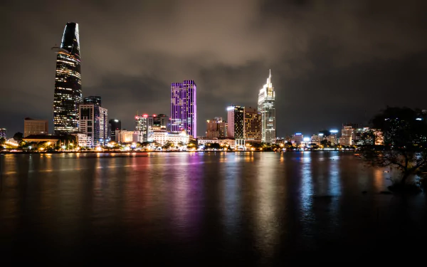 Night man-made skyline of Ho Chi Minh City along the Saigon River, Bitexco Financial Tower among illuminated buildings, lights reflected on water — 8K Ultra HD desktop wallpaper.