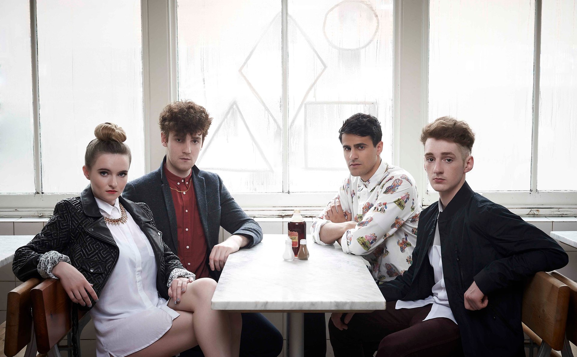 HD desktop wallpaper of Clean Bandit, featuring the band members seated around a table, in a room with bright window light.