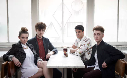 HD desktop wallpaper of Clean Bandit, featuring the band members seated around a table, in a room with bright window light.