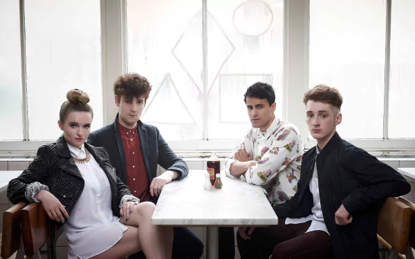 HD desktop wallpaper of Clean Bandit, featuring the band members seated around a table, in a room with bright window light.