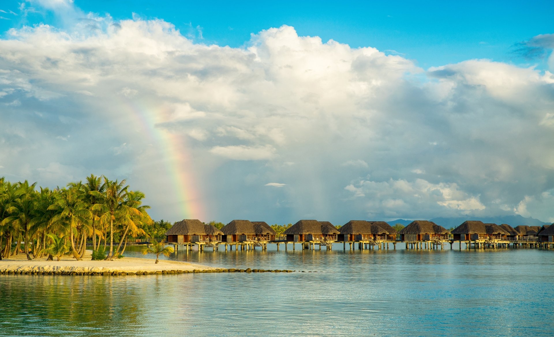 4K Ultra HD image of a tropical bungalow village over calm water, framed by palm trees, a vibrant rainbow, and dynamic clouds under a bright sky.