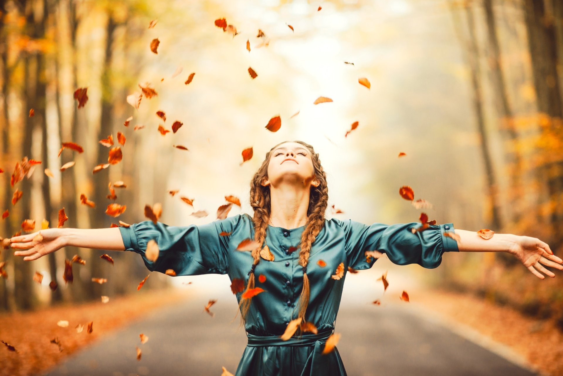 Redheaded woman model with braided hair stands on a tree-lined autumn road, arms outstretched as falling leaves swirl; warm, moody depth-of-field HD desktop wallpaper.