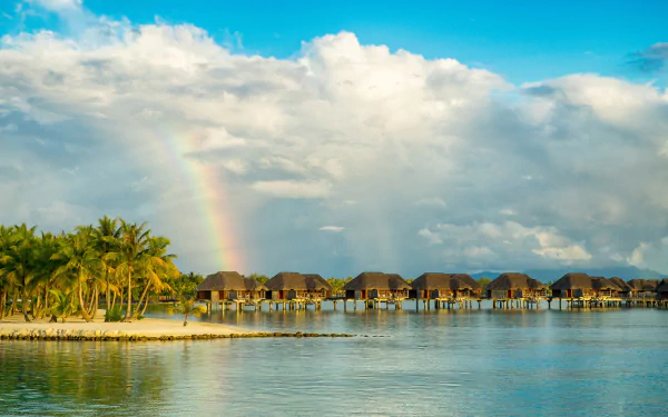 4K Ultra HD image of a tropical bungalow village over calm water, framed by palm trees, a vibrant rainbow, and dynamic clouds under a bright sky.
