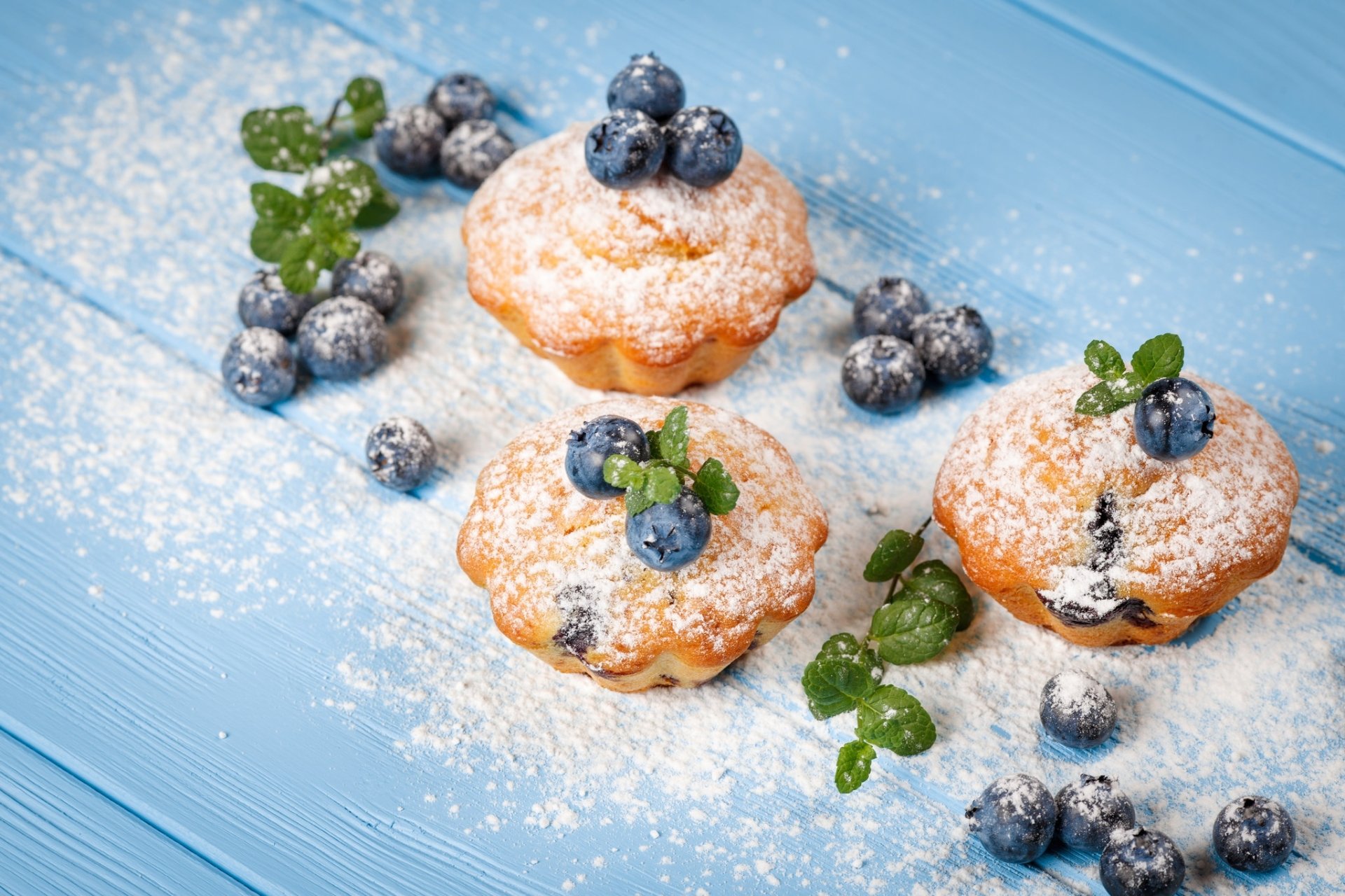 HD desktop wallpaper featuring blueberry Madeleines dusted with powdered sugar on a blue wooden surface, accented with fresh blueberries and green mint leaves.