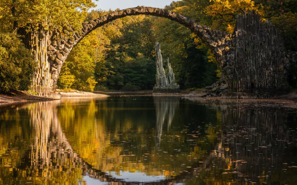 HD desktop wallpaper of the Devil's Bridge, reflecting on a serene river surrounded by lush, autumn foliage.
