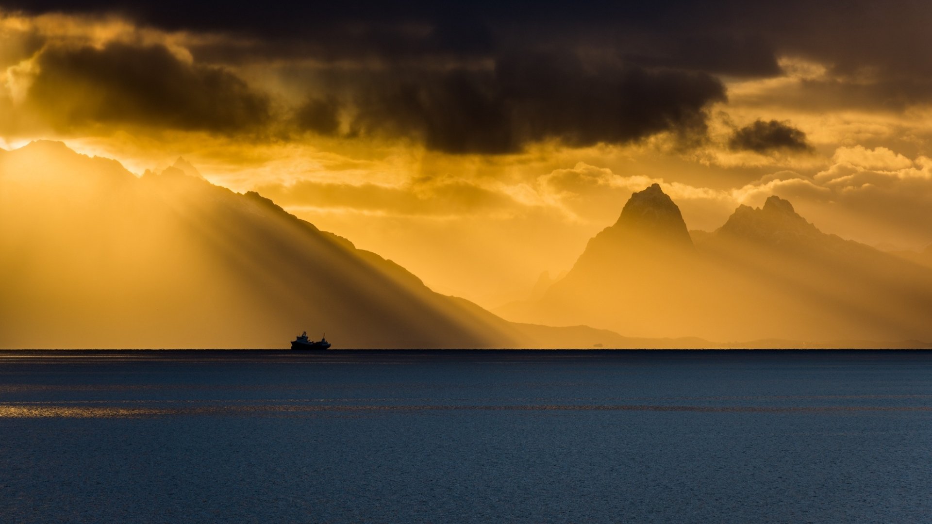 HD wallpaper of a serene coastline at sunset, featuring a ship on the ocean, majestic mountains, and dramatic sunbeams piercing through clouds.