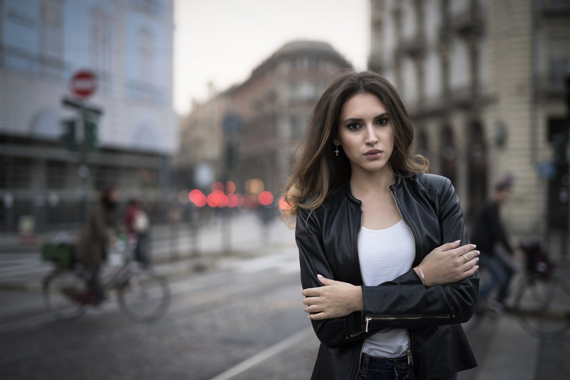Brunette woman with brown eyes in a leather jacket, arms crossed on a blurred city street — shallow depth of field; 2K Quad HD PC desktop wallpaper.