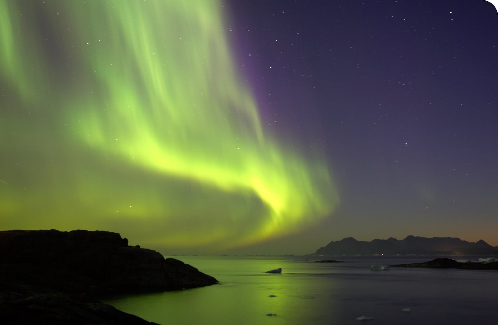 A stunning HD desktop wallpaper showcasing the ocean under the vibrant green and purple hues of the aurora borealis, illuminating the night sky and surrounding mountains.