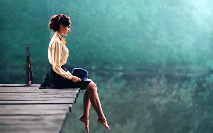 A brunette woman model sits barefoot on a wooden dock, holding a hat, with a soft depth of field background creating a calm and moody HD desktop wallpaper.