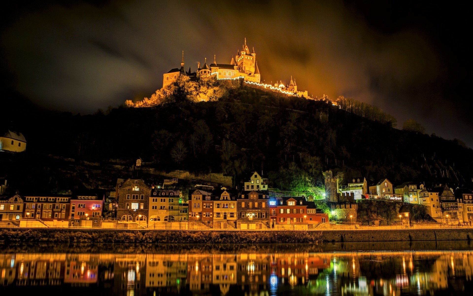 HD desktop wallpaper showing a vibrant cityscape at night with illuminated buildings and a castle reflecting on a calm lake under a cloudy sky.