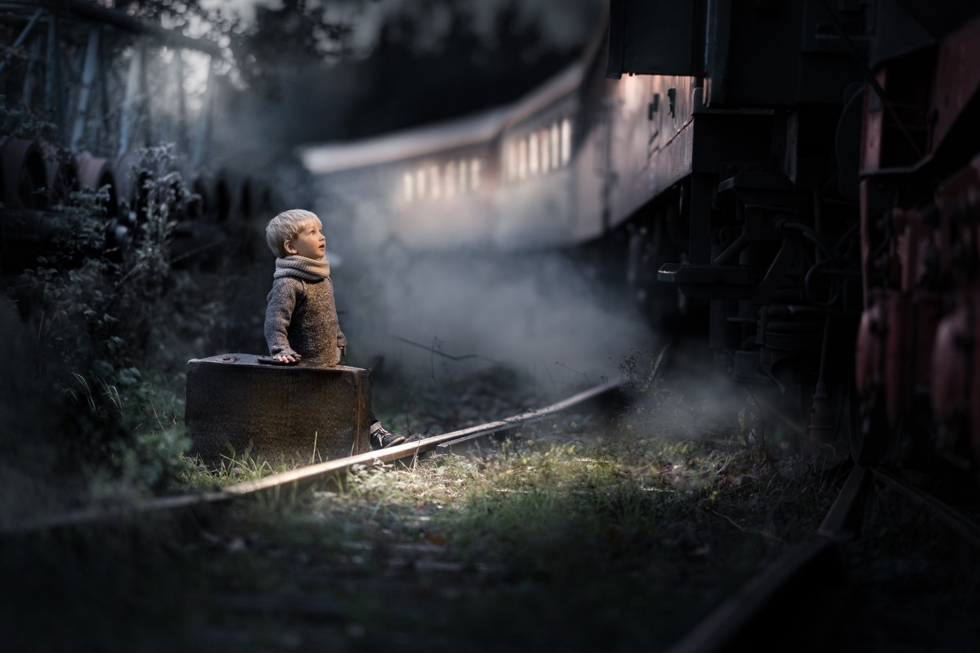 A little boy in a coat sits on a suitcase by the railroad tracks at night, with a train approaching in the mist, captured in a moody, HD desktop wallpaper scene.