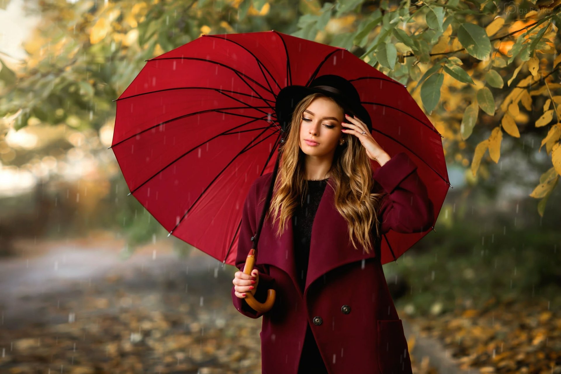 A brunette woman in a coat and hat holds a red umbrella amidst a rainy, autumnal setting, providing a vivid, high-definition desktop wallpaper.