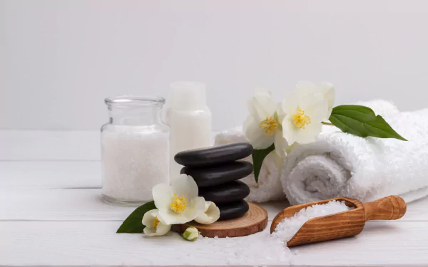 4K Ultra HD still life featuring spa essentials: stacked black stones, white rolled towel, flowers, and jars of bath salts arranged on a white surface.