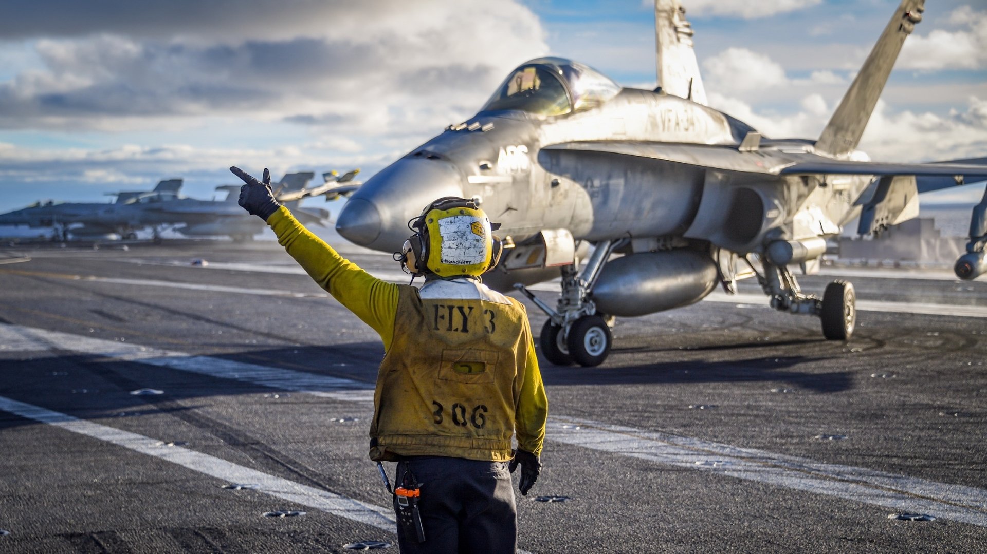 A McDonnell Douglas F/A-18 Hornet jet fighter on an aircraft carrier runway, with a crew member directing it. The image captures a high-definition military scene as a desktop wallpaper and background.