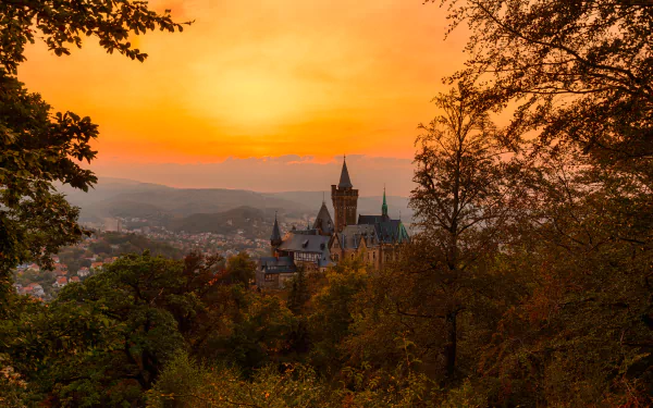 HD desktop wallpaper showing a stunning sunset over a German castle, surrounded by lush trees and a serene landscape. The warm hues of the setting sun create a captivating background.
