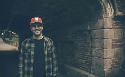 Man in a plaid shirt and red cap smiling under a bridge, HD desktop wallpaper background.