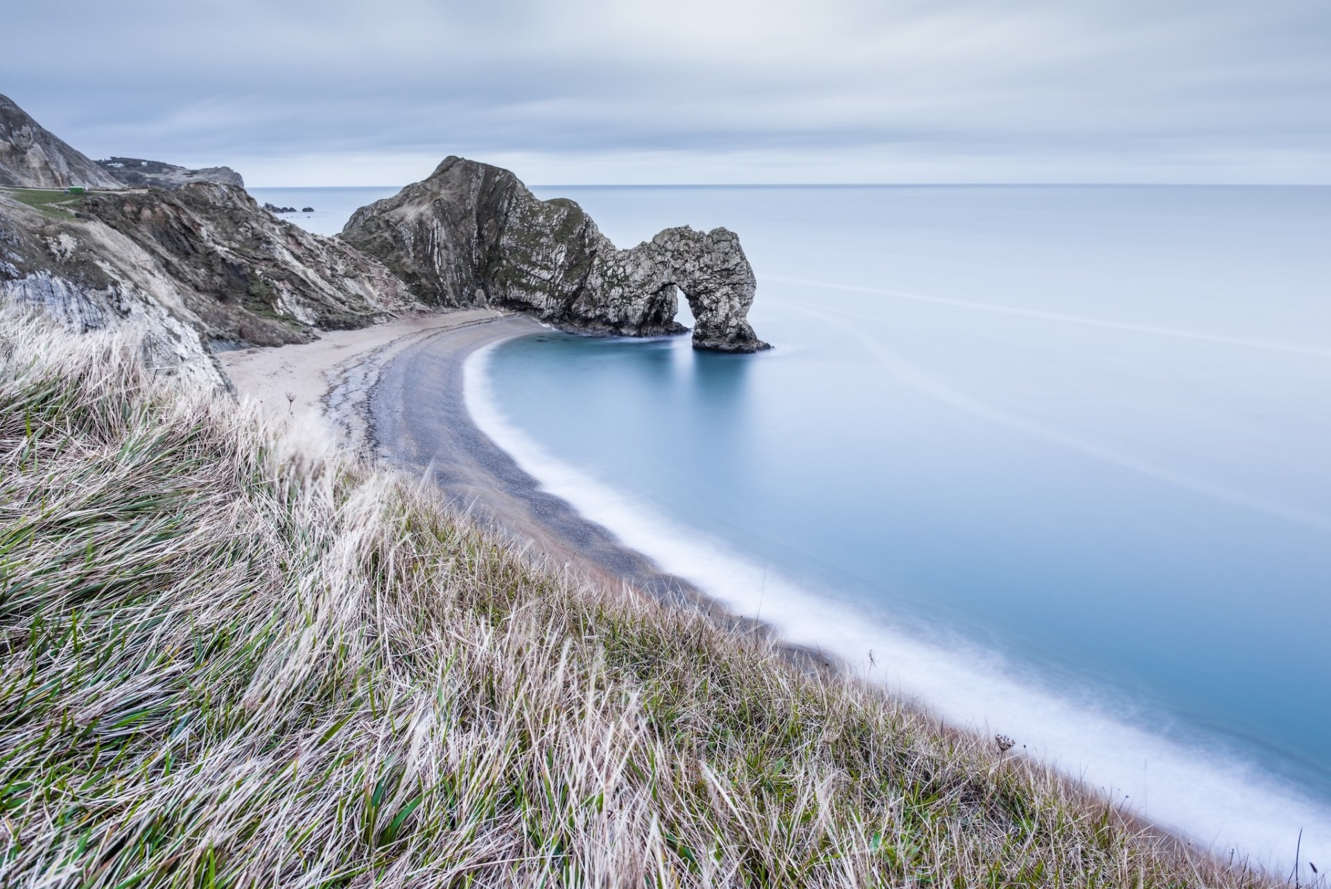 HD desktop wallpaper of Durdle Door’s natural limestone arch along the calm English coast, featuring a serene ocean horizon and beach framed by coastal cliffs and grasses.