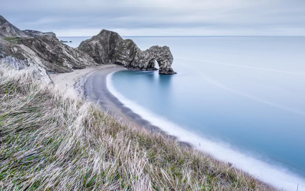 HD desktop wallpaper of Durdle Door’s natural limestone arch along the calm English coast, featuring a serene ocean horizon and beach framed by coastal cliffs and grasses.