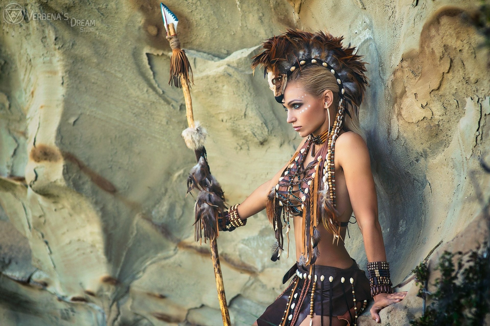 HD PC desktop wallpaper of a woman in intricate tribal cosplay holding a decorated spear, standing against a textured rocky background.