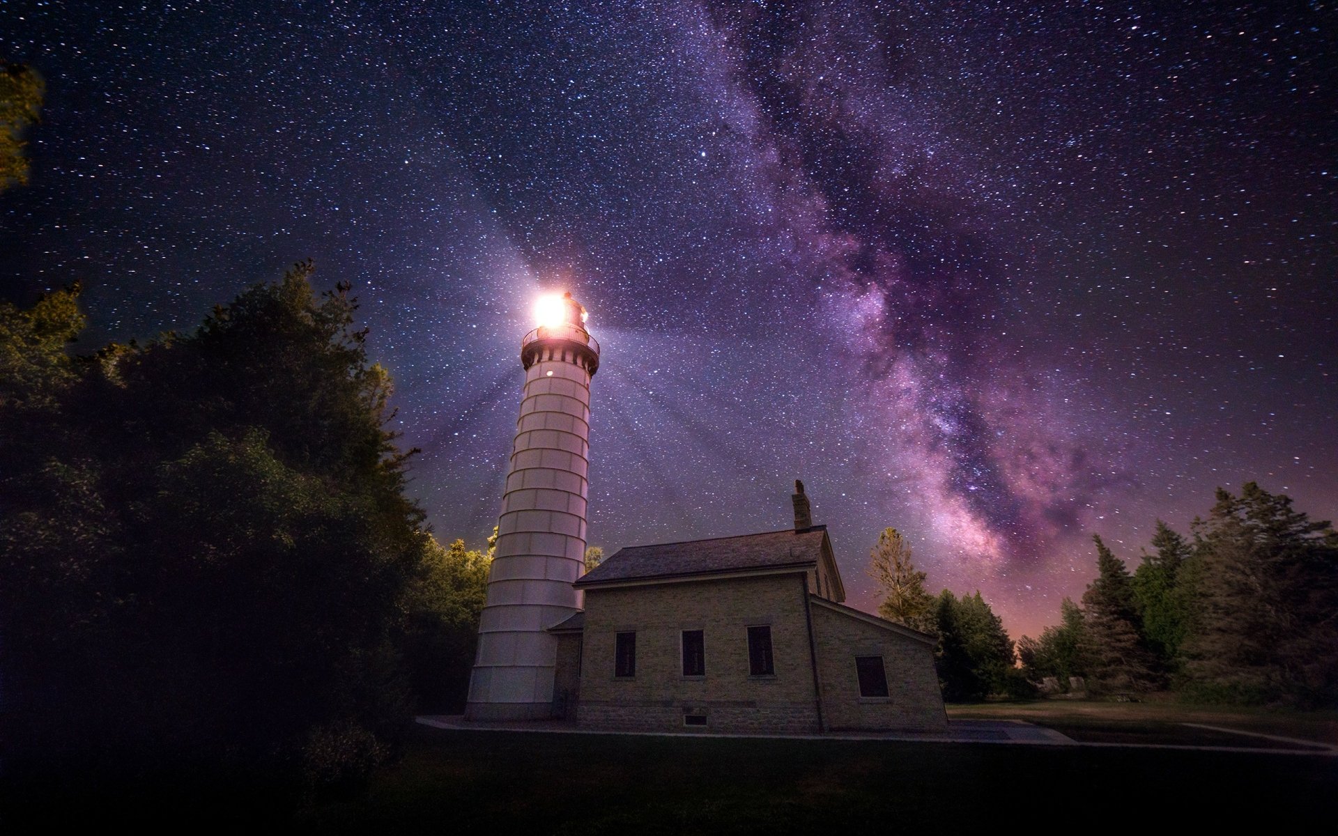A lighthouse illuminated under a vibrant, starry sky with the Milky Way stretching across the night, captured in high-definition for a desktop wallpaper background.