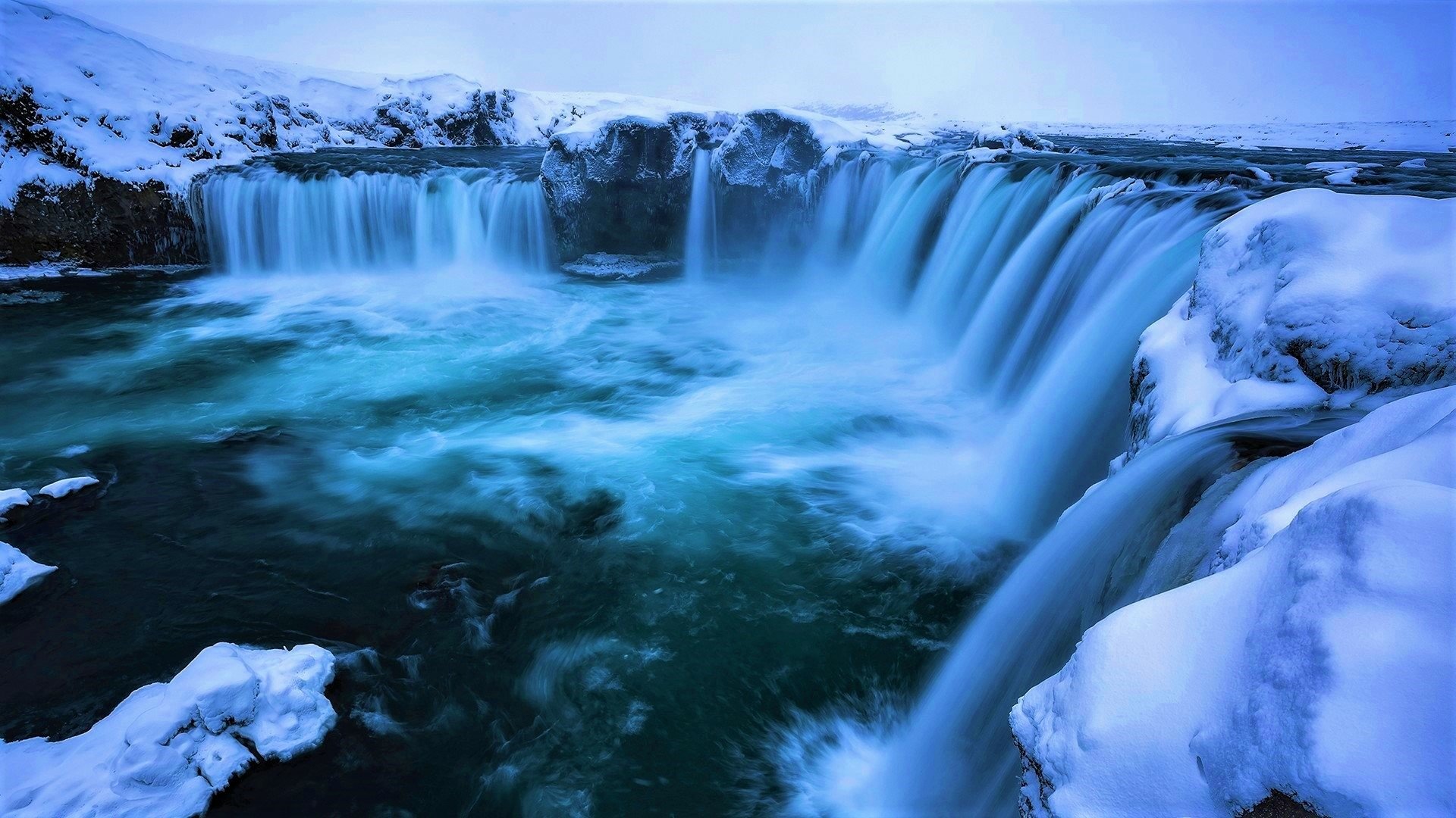 HD desktop wallpaper of Goðafoss waterfall surrounded by snow and icy winter nature under a soft blue sky.