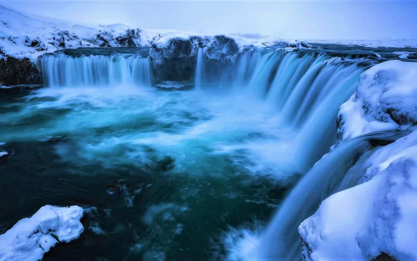 HD desktop wallpaper of Goðafoss waterfall surrounded by snow and icy winter nature under a soft blue sky.