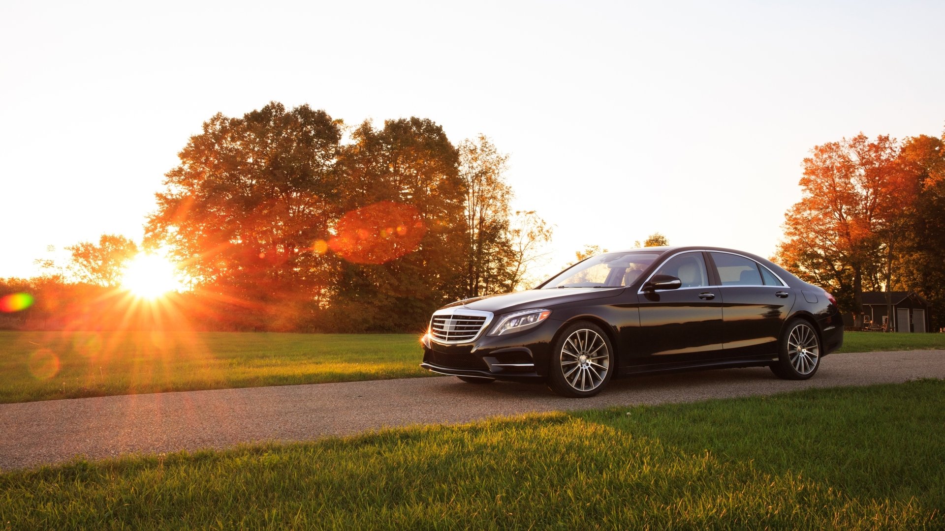 A black Mercedes-Benz S-Class car parked on a road with trees in autumn colors and the sun setting in the background, captured in 4K Ultra HD quality.