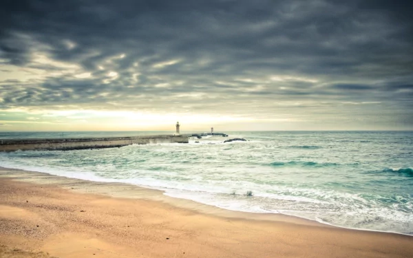 A serene coastline features a lighthouse standing sentinel by the sea, with gentle waves lapping at the sandy shore under a dramatic cloud-filled sky. A stunning nature landscape.