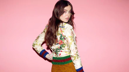 An HD desktop wallpaper featuring American actress and singer Hailee Steinfeld with brunette hair, posing against a pink background. She wears a floral top and looks over her shoulder.