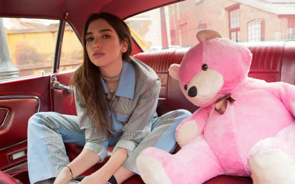 A brunette singer sits in a retro car next to a large pink teddy bear, embodying a playful and vibrant music-inspired vibe in this HD desktop wallpaper.